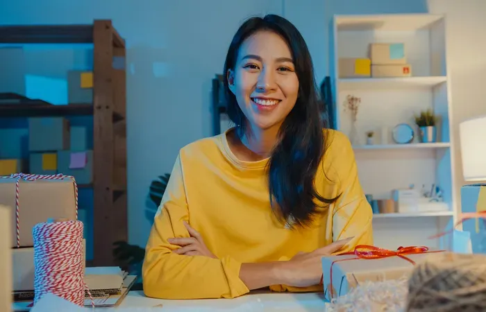 Smiling woman in yellow sweater sitting at desk packaging handmade products in cozy small business workspace